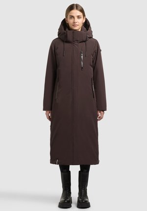 Vrouw staat naar voren gericht, draagt een lange donkerbruine winterjas met capuchon, ritssluitbare zakken en zwarte laarzen tegen een effen achtergrond.