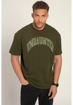 Camiseta de algodón verde oliva con "UNDAUNTED" impreso en un diseño de arco en verde más claro. Con mangas cortas y cuello redondo, combinada con pantalones oscuros.