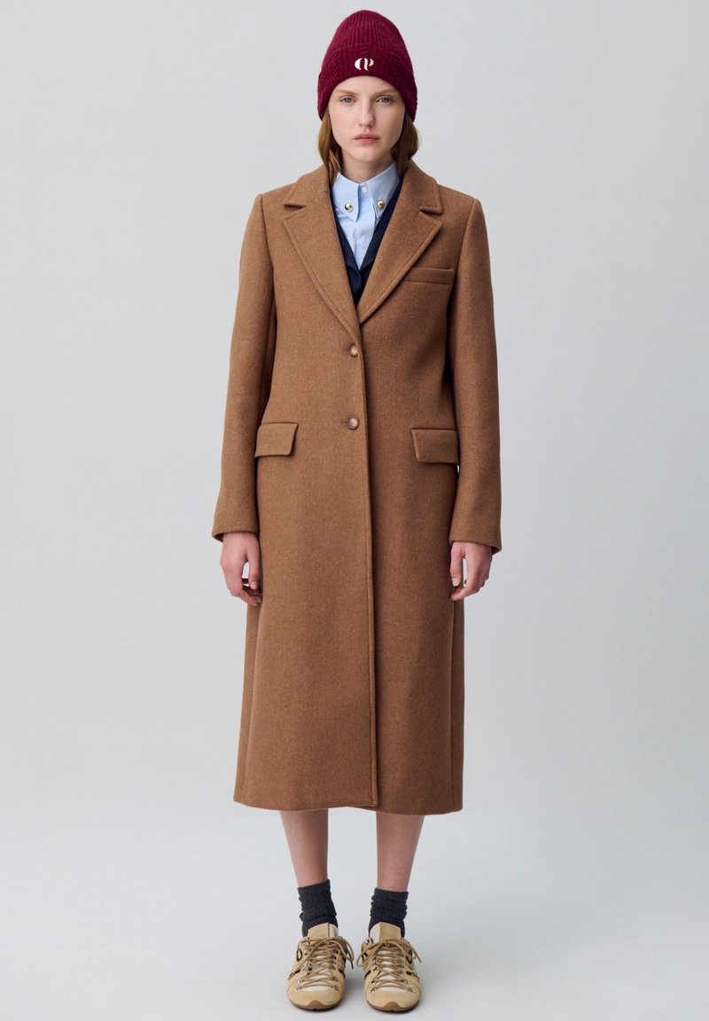 Cappotto marrone in lana con design su misura, due tasche frontali e chiusura con un solo bottone. Abbinato a una camicia blu e un berretto a maglia bordeaux.