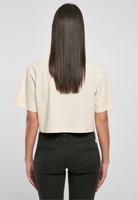 Persona con lunghi capelli castani lisci, che indossa un crop top beige a maniche corte e jeans neri a vita alta, in piedi con le spalle rivolte verso la fotocamera su uno sfondo semplice.