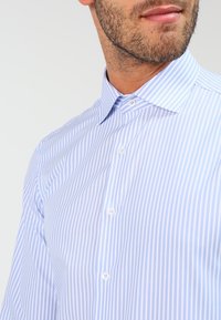 Man in een lichtblauw en wit verticaal gestreept overhemd met kraag, die zijn ondergezicht en nek laat zien met een lichte gezichtsbeharing.