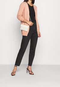 Svetle růžový kardigan s dlouhými rukávy z měkkého úpletu, doplněný o černé džíny s vysokým pasem a černé sandály na podpatku s pásky. Detail bílého kabelky s korálky.