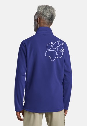 Mann trägt eine blaue Fleecejacke mit einem weißen Pfotenabdruck auf dem oberen Rücken und steht vor einem schlichten Hintergrund.