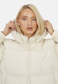 Kurtka puchowa w kolorze off-white z teksturowanym, polarowym kapturem i zapięciem na zamek. Posiada boczne kieszenie oraz elastyczne mankiety.
