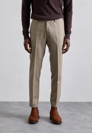 Mężczyzna w beżowych, dopasowanych spodniach, brązowych zamszowych butach i ciemnobrązowej bluzce z długim rękawem stoi na szarej podłodze.