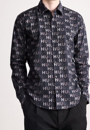 Mężczyzna ubrany w czarną koszulę na guziki z nadrukiem powtarzającego się jasnoszarego napisu „HUG”, w połączeniu z czarnymi spodniami, ręce w kieszeniach.