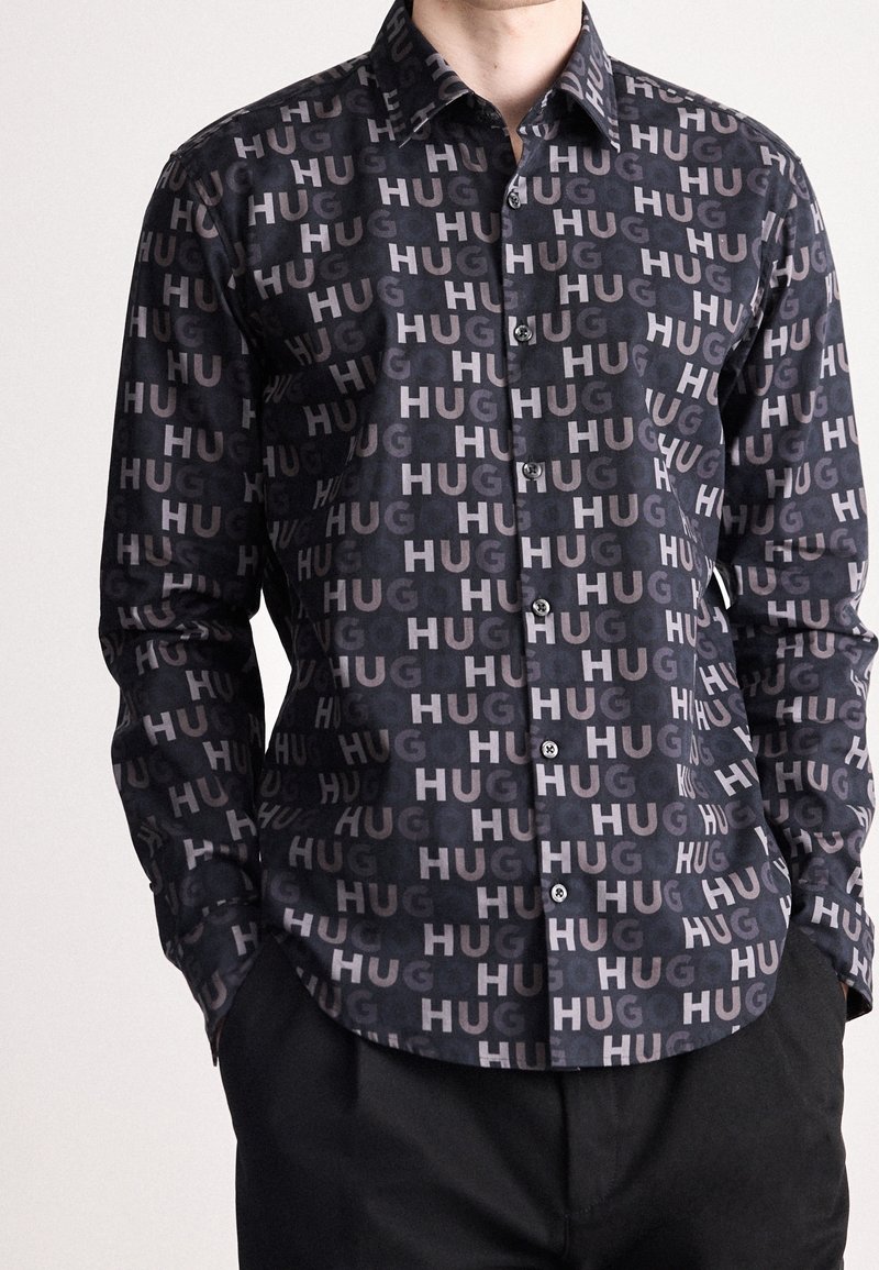 Homme portant une chemise noire à boutons imprimée de manière répétée avec le texte "HUG" en gris clair, associée à un pantalon noir, mains dans les poches.