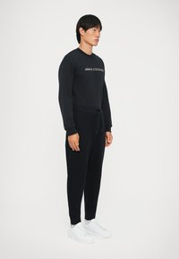 Melnā sporta jakas un bikses komplekts, kuram ir aukliņa viduklī, ribotie gadījumi un "ARMANI EXCHANGE" logo uz krūtīm.