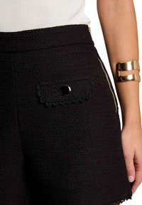 Shorts noirs texturés avec une poche festonnée dotée d'un bouton noir et une fermeture à glissière sur le côté visible. Manchon en or métallique au poignet.