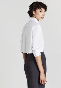 Weiße cropped Bluse mit einer Rückenfalte und aufgerollten Ärmeln, kombiniert mit einem dunkelgrauen, taillierten Rock, der eine subtile Textur aufweist.