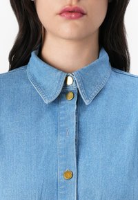 Camisa de denim azul claro com colarinho clássico, botões dourados e detalhes de costura visíveis. Textura suave e design casual.