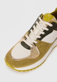 Olive green and beige sneaker with smooth and textured materials, gold accents, adjustable laces, and a thick white sole with yellow detailing.