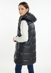 Sort polstret vest med hætte, der har en blank overflade, sidelommer og en høj krave. Modellen har det på over en hvid sweater.