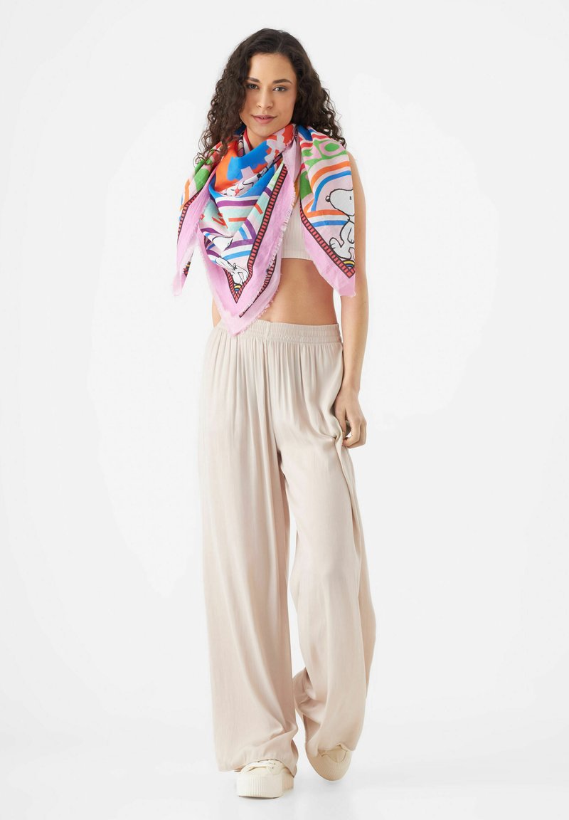 Écharpe en soie multicolore avec des motifs amusants, drapée autour du cou. Le modèle porte un pantalon large fluide beige et des baskets crème.