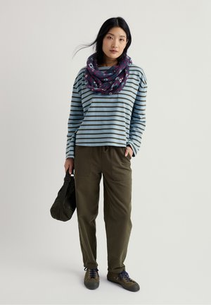 Žena koja nosi plavu blago prugastu majicu dugih rukava, maslinasto zelene hlače, cvjetni šal i maslinaste tenisice, držeći tamno zelenu torbu.