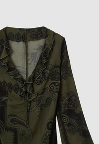 Blouse vert olive avec un motif paisley noir complexe, encolure froncée et détail noué à l'avant. Manches longues en tissu texturé.