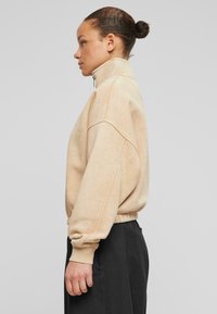 Beige gyapjú pulóver magas gallérral, eső vállakkal és rugalmas mandzsettákkal; sötét nadrággal párosítva, amely laza szabású és minimális részletezéssel rendelkezik.