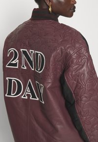 Bordová kožená bomber bunda s vyšívaným kvetinovým dizajnom a veľkým nápisom "2ND DAY" v čiernej a bielej farbe. Čierne rukávy s rebrovaným golierom.