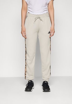 Person iført beige Adidas joggers med leopardprintstriber på siderne, parret med sorte Adidas sneakers og en hvid skjorte.