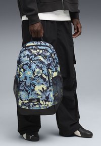 Eine Person hält einen blau-gelben, blumigen Puma-Rucksack, trägt schwarze Hosen, schwarz-weiße Puma-Schuhe und eine schwarze Jacke über einem weißen Shirt.
