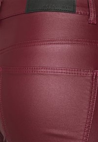 Burgundowe spodnie z ekoskóry z widocznymi szwami, podkreślonymi tylnymi kieszeniami oraz czarną metką z napisem "NOISY MAY" na tylnej części pasa.