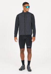 Svart lättviktsväst med dragkedjeförsedd bröstficka, långa svarta ärmar, svarta cykelshorts, svarta strumpor och svarta cykelskor. Blå solglasögon.