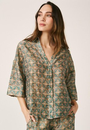 Mujer vistiendo una camisa holgada de botones con estampado en tonos teal y beige, con mangas tres cuartos, y pantalones a juego, de pie con las manos en los bolsillos.