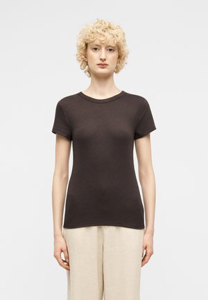 Γυναίκα με κοντά σγουρά ξανθά μαλλιά, που φοράει εφαρμοστό σκούρο καφέ κοντομάνικο πουκάμισο και φαρδιά μπεζ παντελόνια, στέκεται μπροστά σε λευκό φόντο.