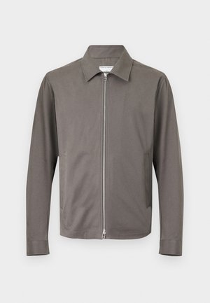 Ung mand med krøllet hår iført grå jakke, mørk skjorte, blå jeans, hvide sneakers, som holder en brun lædertaske, stående mod en hvid baggrund.