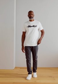 Valge puuvillane T-särk, millel on "Antony Morato" tekst, hall slim-fit püksid ja valged tossud. Puitpõrand ja lihtne taust.