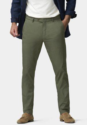 Man draagt olijfgroene slim-fit broek, tan suède loafers met kwastjes, wit overhemd en een marineblauw colbert met handen in de zakken.