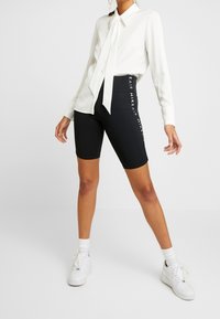 Camicia bianca a maniche lunghe con dettaglio della cravatta, abbinata a pantaloni corti neri da biker con testo bianco sui lati. Indossata con sneakers bianche.