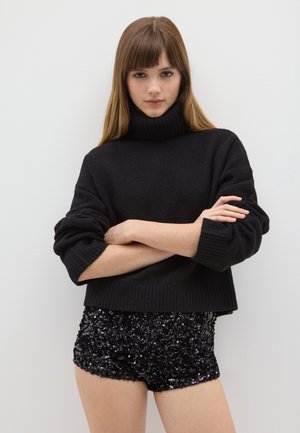 Pull à col roulé noir en tricot doux avec des accents côtelés, associé à un short noir à sequins. Le modèle pose les bras croisés devant un fond blanc.