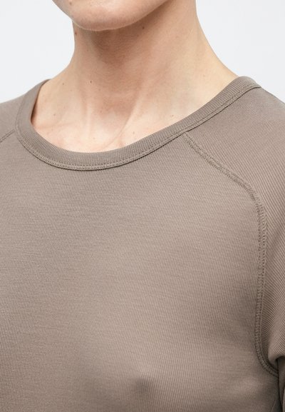 Tuvplāns ar personu, kas valkā pelēkbrūnu ribotu auduma kreklu ar redzamu šuvumu pie apkakles un pleca.