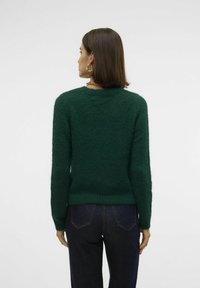 Zelena, strukturirana jopica z dolgimi rokavi in okroglim izrezom, povezana s temno jeans hlačami. Mehka, puhasta tkanina in prilegajoča zasnova.
