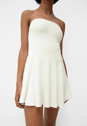 Mujer con un vestido blanco sin tirantes, texturizado, con cuerpo ceñido y falda acampanada sobre un fondo blanco liso.