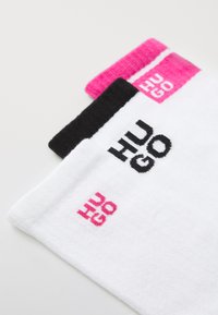 Három pár zokni: fehér, fekete és rózsaszín díszítéssel. Minden zoknin a "HUGO" felirat kontrasztos színekben jelenik meg. Puha, szőtt anyagú textúra.