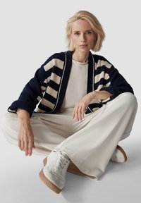 Gestreifter, navy- und cremefarbener Strickcardigan über einem hellen Oberteil, kombiniert mit cremefarbenen, weiten Hosen und weißen Sneakers mit einer hellbraunen Sohle.