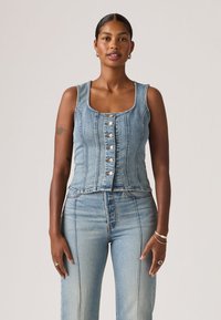 Γυναίκα με σκουρόχρωμο δέρμα και μαλλιά σε κότσο, φορεί στενόμηκη τζιν γιλέκο με κουμπιά χωρίς μανίκια και τζιν παντελόνι ανοιχτού μπλε χρώματος με ψηλή μέση, στέκεται μπροστά σε ουδέτερο φόντο.