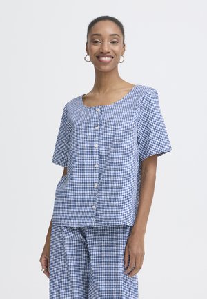 Lachende vrouw die een blauw-wit geruit, kort mouwtjes overhemd met knopen draagt en bijpassende wijde broek, staand voor een neutrale achtergrond.