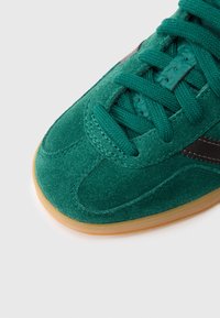 Zeleni semiš superga z teksturirano površino, črnimi poudarki in gumijastim podplatom; ima okroglo konico in ravne vezalke.