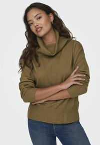 Pull à col montant vert olive avec des accents côtelés, manches longues et coupe décontractée, associé à un jean en denim bleu. Tissu doux et texturé.