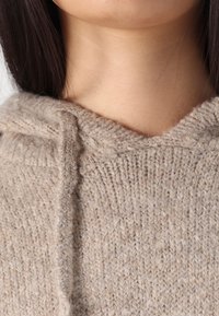Felpa beige lavorata a maglia con tessuto testurizzato, caratterizzata da un collo a V e dettagli di cucitura visibili lungo il bordo e il cappuccio.