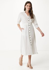 Abito bianco con bottoni, con vita cintata, realizzato in tessuto leggero. Presenta maniche lunghe, tasche laterali e orlo midi.