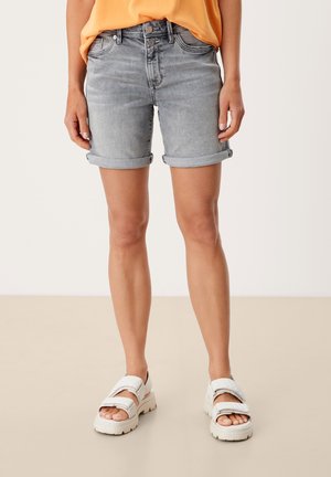 Person iført opsmøgede grå cowboyshorts, orange top og hvide chunky sandaler, stående på en beige gulv med en ensfarvet baggrund.