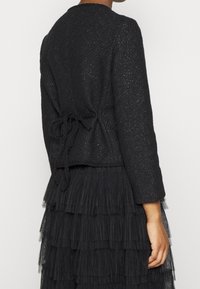 Černá strukturovaná bunda s tkaným vzorem a stahováním v pase, doplněná vrstvenými černými třásňovými šaty.