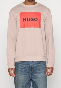 Sudadera color beige claro con un gráfico rectangular rojo que presenta la palabra "HUGO" en negro. Cuello y puños acanalados. Llevada con jeans azules.