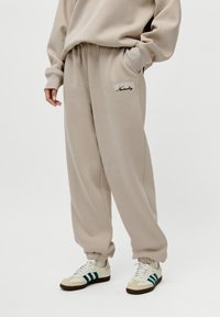 Beige joggebukser med elastisk linning og mansjetter, med brodert "MODERN Neutrality" logo på fronten. Myk, glatt stoff.