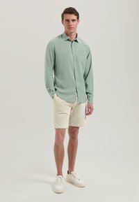 Cămașă lungă, verde deschis, cu mâneci lungi și buzunar frontal, asociată cu pantaloni scurți texturați bej și adidași albi. Outfit simplu și casual.