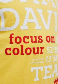 Gelber Stoff mit erhabenem weißem und rotem Text mit der Aufschrift "focus on colour" sowie teilweise sichtbaren Wörtern, Koordinaten und "Authentic Trademark".